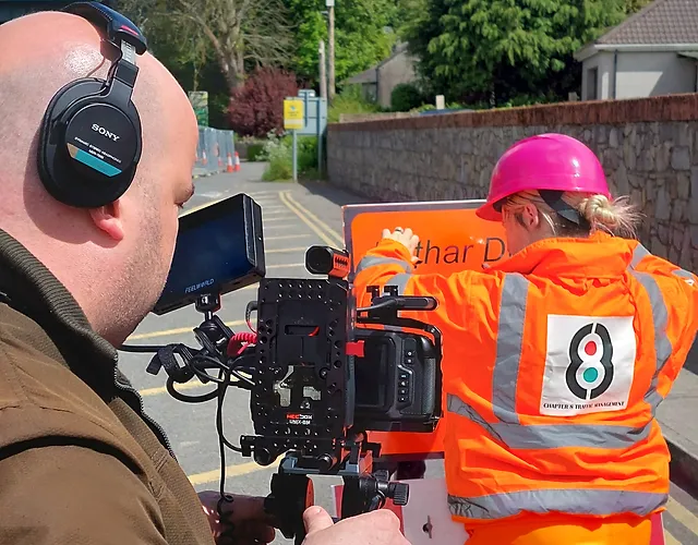 Professional camera man films female construction worker with pink hard hat working on a road project in the sun